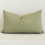 Sage solid-colour cushion positioned in front of a brick wall. It has rectangle dimensions and is made from an outdoor material.