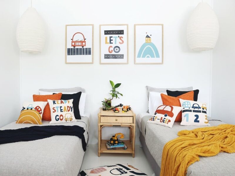 Kids bedroom with car kids cushions on twin beds with a throw blanket. On the wall are three kids wall prints that match the cushions
