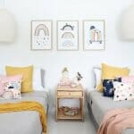 Kids cushions with rainbow designs on two single grey beds with a pink throw blanket. On the wall are three rainbow wall prints matching the cushions