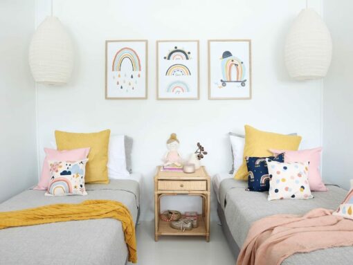 Kids cushions with rainbow designs on two single grey beds with a pink throw blanket. On the wall are three rainbow wall prints matching the cushions