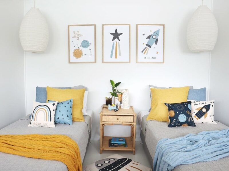 Kids cushions with moons and stars designs on twin grey beds with a light blue throw blanket. On the wall are three stars and moon wall prints matching the cushions