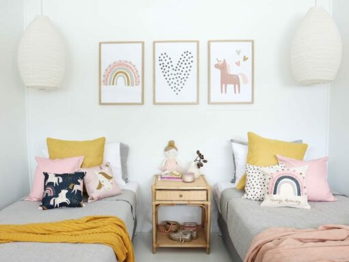 Kids cushions with unicorn prints in pink sitting on two beds with throw blankets on them. On the wall are matching unicorn kids wall prints.