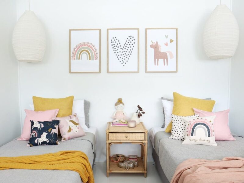 Kids cushions with unicorn prints in pink sitting on two beds with throw blankets on them. On the wall are matching unicorn kids wall prints.