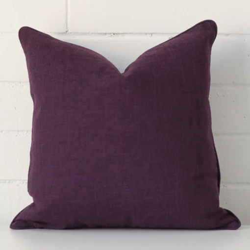 A square cushion in a delightful plum tone rests against a white wall. The linen material appears to be of exceptional quality.