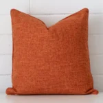 A brick wall that has a rust cushion cover positioned in front of it. It has an exquisite linen material and a lovely square shape.