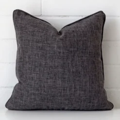 Grey cushion leans elegantly against a brick wall. It has been crafted from a high quality linen material and has a square shape.