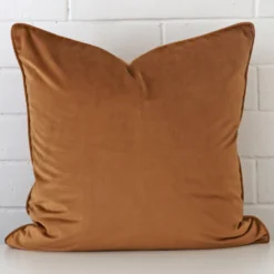 A velvet large cushion cover that is shown vertically against a brick wall. It has a wonderful bronze colour.