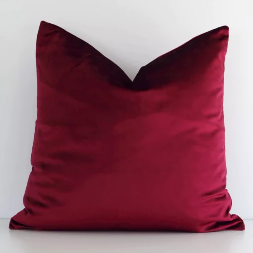 A white wall that has a solid coloured burgundy cushion cover positioned in front of it. It has an exquisite velvet material and a lovely square shape.