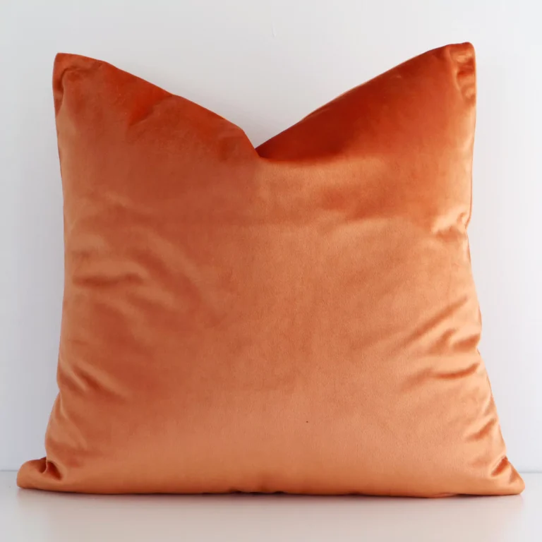 A square solid colour cushion in a delightful burnt orange tone rests against a white wall. The velvet material appears to be of exceptional quality.