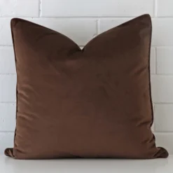 Chocolate brown cushion leans elegantly against a brick wall. It has been crafted from a high quality velvet material and has a large size.
