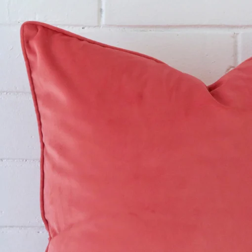 Enlarged shot of the corner of this large cushion cover in coral colour is shown against a brick wall. The image shows the quality and craftsmanship of the velvet material.
