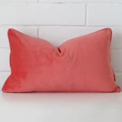 A velvet rectangle cushion cover that is shown vertically against a brick wall. It has a wonderful coral colour.