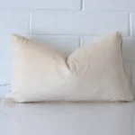 A rectangle cream cushion positioned in front of white brickwork. It has a velvet fabric.