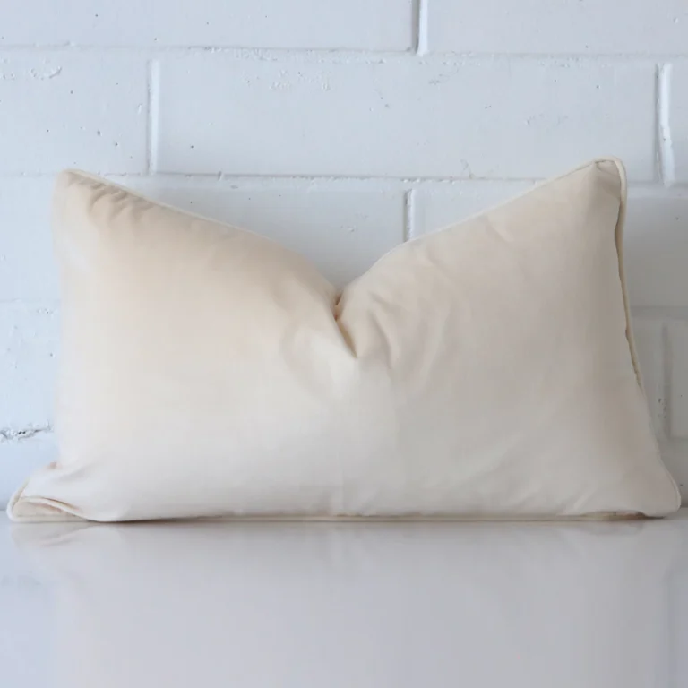 A rectangle cream cushion positioned in front of white brickwork. It has a velvet fabric.