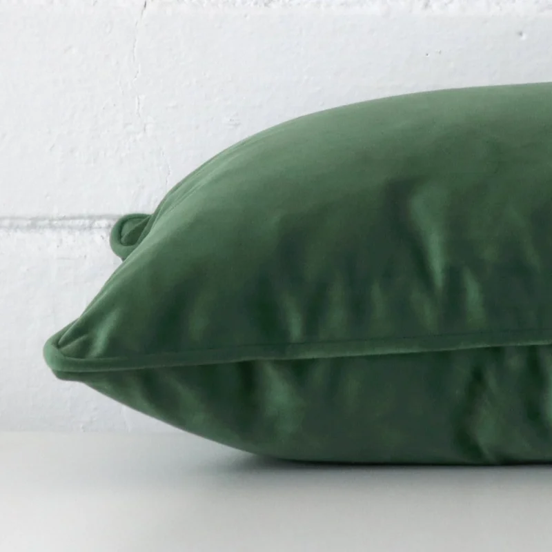 A brick wall that has a dark sage cushion cover positioned in front of it. It has an exquisite velvet material and a lovely rectangle shape.