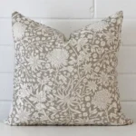 A linen square cushion cover that has a unique floral design is shown vertically against a brick wall. It has a wonderful grey colour.