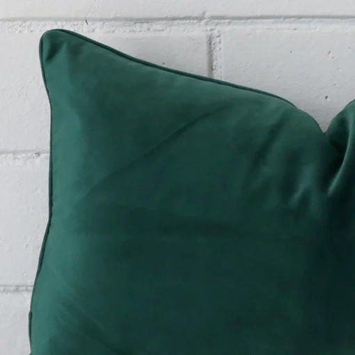 Enlarged shot of the corner of this rectangle cushion cover in emerald green colour is shown against a brick wall. The image shows the quality and craftsmanship of the velvet material.