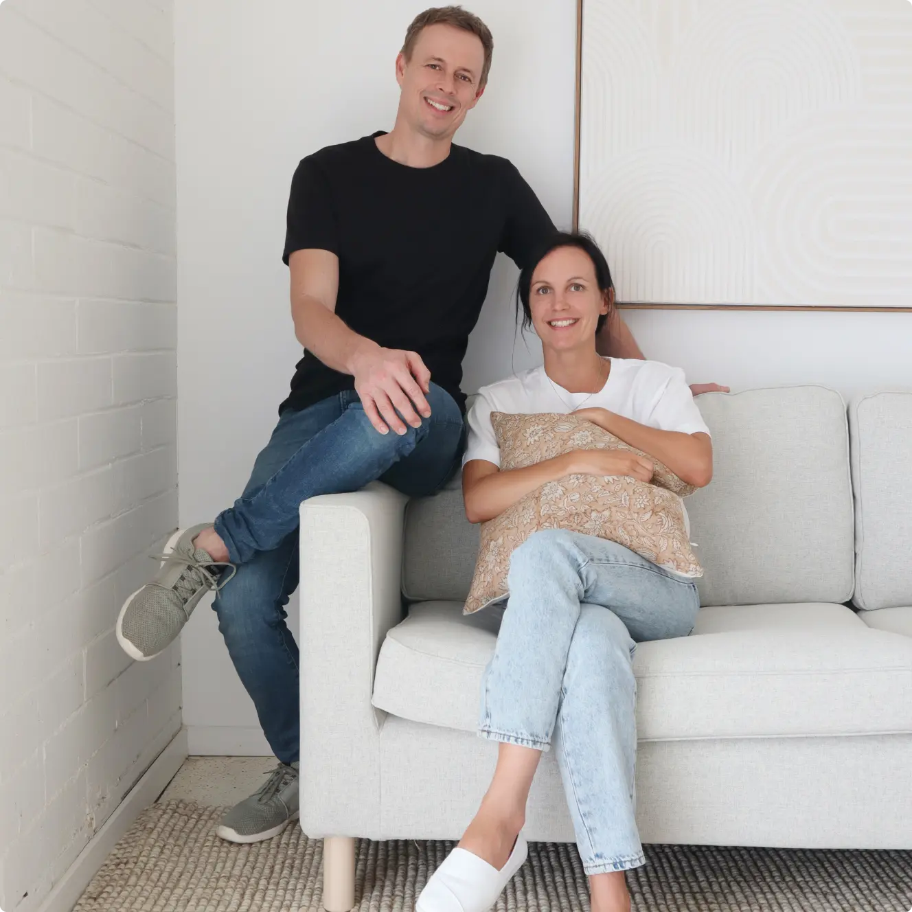 The founders, a couple, are sitting on a couch with artwork on the wall.