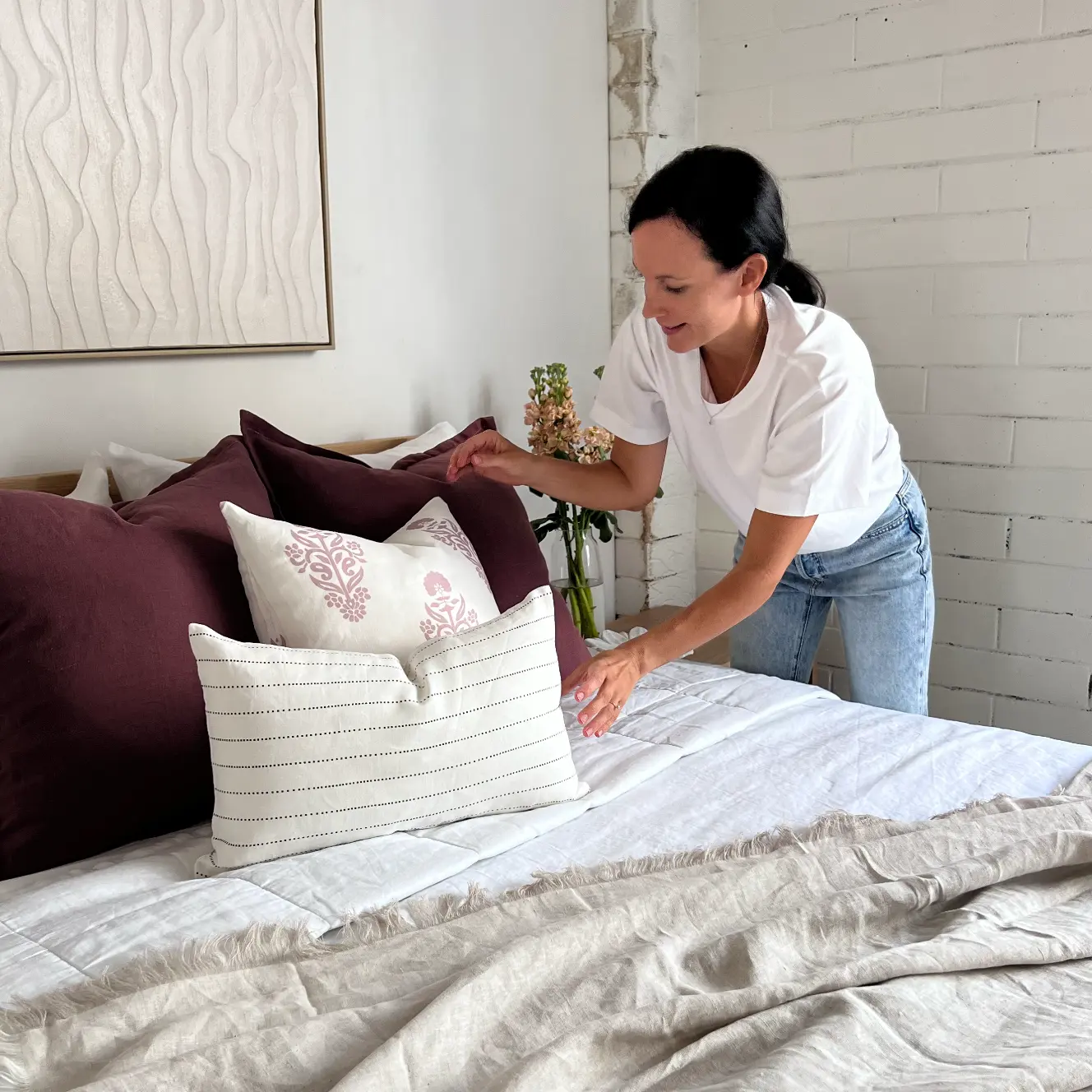 A lady is styling some cushions in a light airy bedroom.