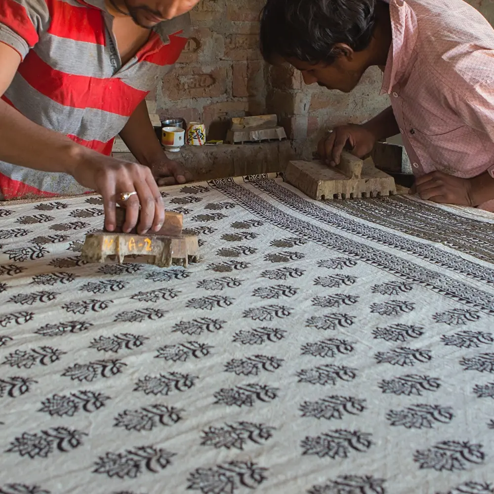 A large roll of fabric is being carefully block printed by two artisans.