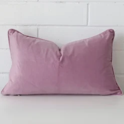 Lavender cushion positioned in front of a brick wall. It has rectangle dimensions and is made from a velvet material.