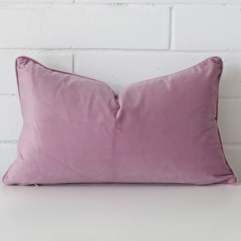 lavender-rectangular-velvet-cushion-cover-30cm-x-50cm-sc498-30c Lavender cushion positioned in front of a brick wall. It has rectangle dimensions and is made from a velvet material.
