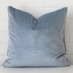 Here a light blue velvet cushion is shown styled against a white wall. It has a large size.