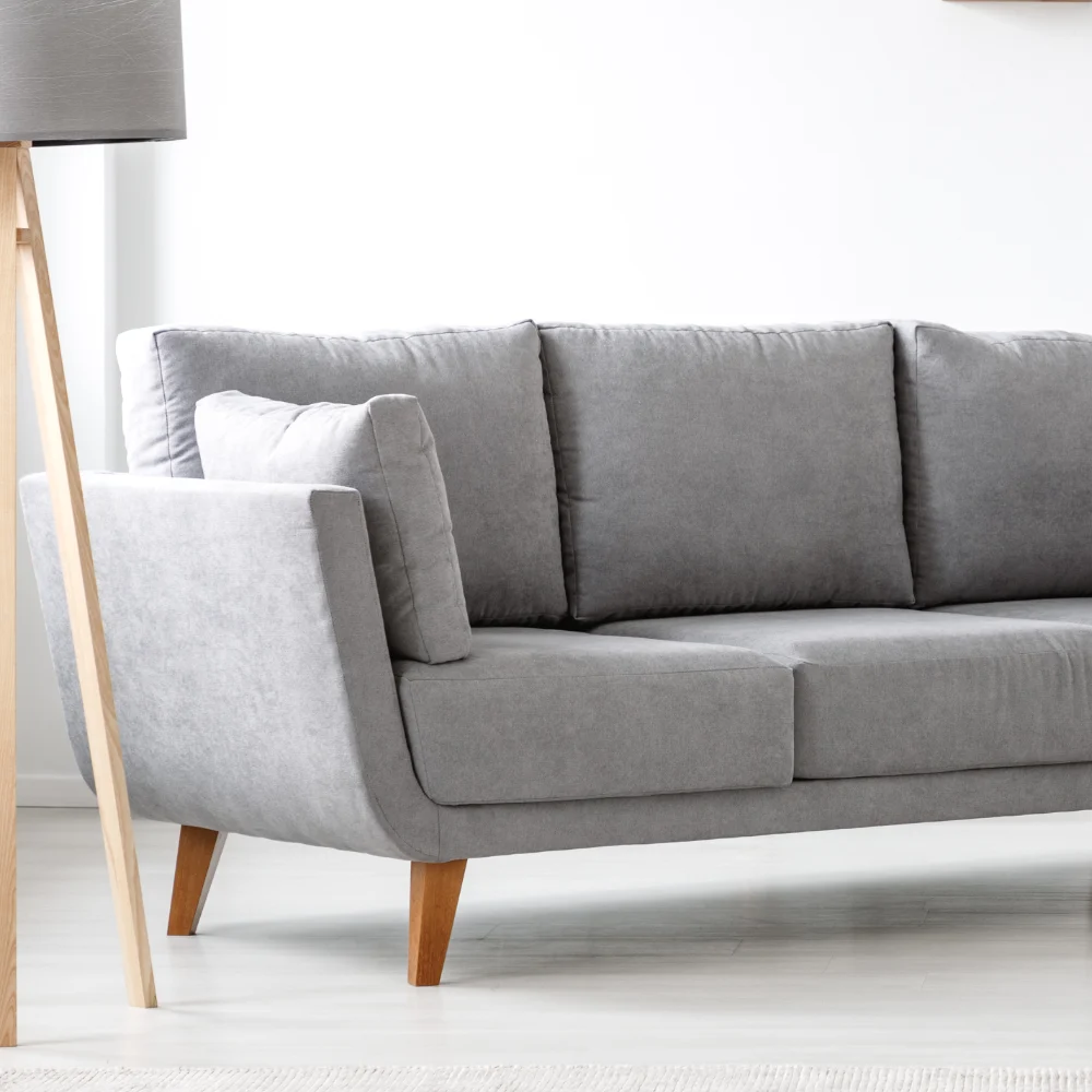 A light filled room has a floor lamp standing next to an empty grey couch. Perfect blank canvas for cushions for a green sofa.