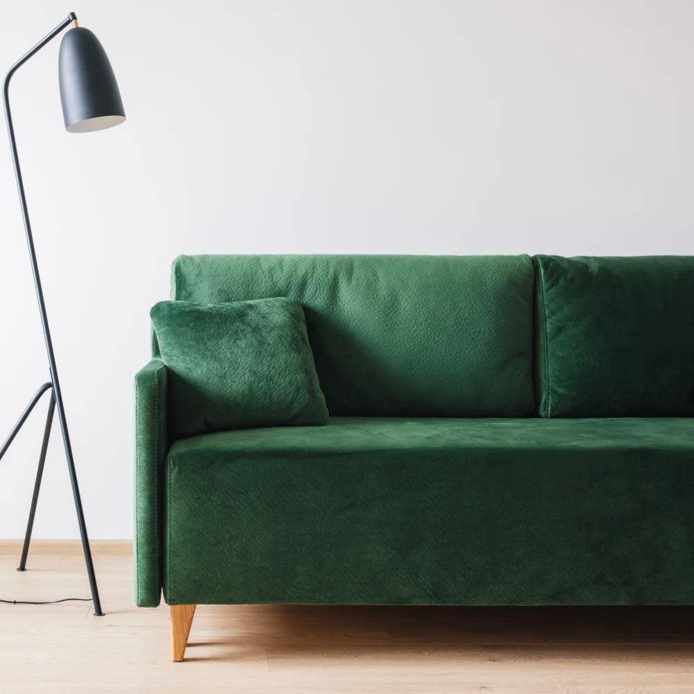 A light filled room has a floor lamp next to an empty green couch. Perfect blank canvas for cushions for a green sofa.