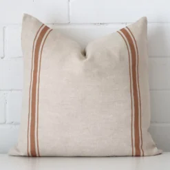 A pretty striped linen cushion cover is shown against a brick wall. It features a square shape and a terracotta colour finish.