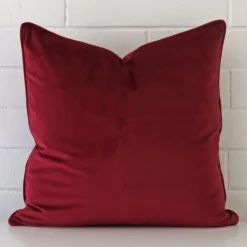 A large cushion in a delightful maroon tone rests against a white wall. The velvet material appears to be of exceptional quality.