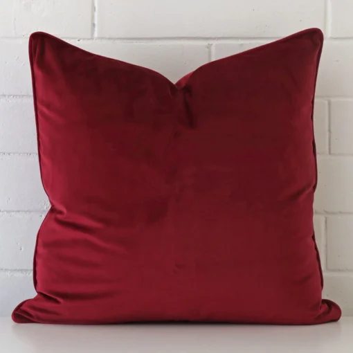 A large cushion in a delightful maroon tone rests against a white wall. The velvet material appears to be of exceptional quality.