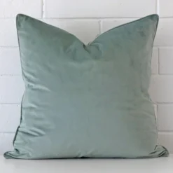 Mint blue cushion positioned in front of a brick wall. It has large dimensions and is made from a velvet material.