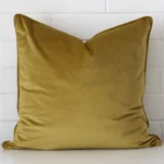 A brick wall that has a mustard cushion cover positioned in front of it. It has an exquisite velvet material and a lovely large shape.