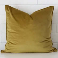 A brick wall that has a mustard cushion cover positioned in front of it. It has an exquisite velvet material and a lovely large shape.