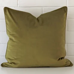 A pretty velvet cushion cover is shown against a brick wall. It features a large shape and an olive colour finish.