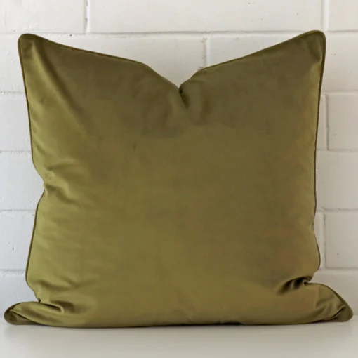 A pretty velvet cushion cover is shown against a brick wall. It features a large shape and an olive colour finish.
