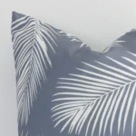 Enlarged shot of the corner of this square palm printed cushion cover in grey colour is shown against a white wall. The image shows the quality and craftsmanship of the outdoor material.