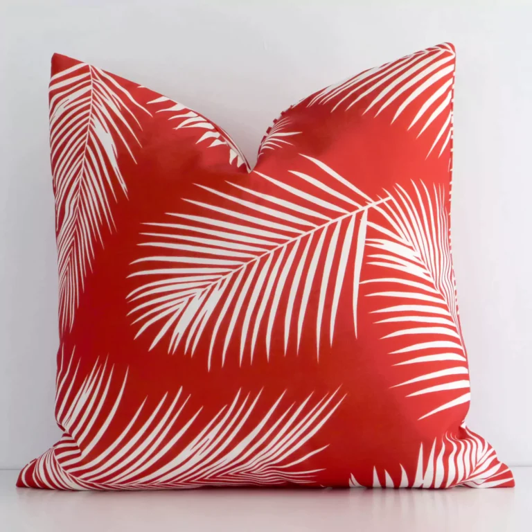 Red palm printed cushion leans elegantly against a white wall. It has been crafted from a high quality outdoor material and has a square shape.