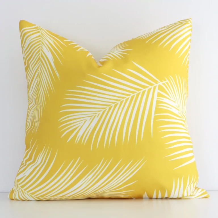 A squate palm printed cushion in a delightful yellow tone rests against a white wall. The outdoor material appears to be of exceptional quality.