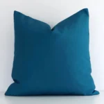 Teal outdoor cushion cover features prominently against a white wall. It is a square design and has a charming solid colour design.