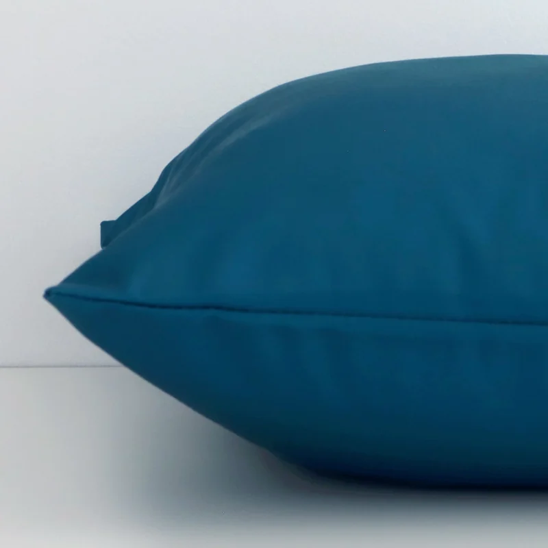 Square solid-coloured cushion cover in teal colour sitting flat. The sideways viewpoint shows the seams of the outdoor material.