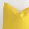 Enlarged shot of the corner of this square solid-coloured cushion cover in yellow colour is shown against a brick wall. The image shows the quality and craftsmanship of the outdoor material.
