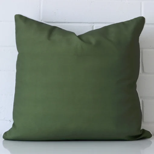 Olive green cushion leans elegantly against a brick wall. It has been crafted from a high quality outdoor material and has a large size.