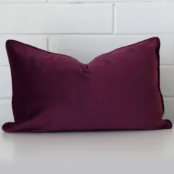 A rectangle cushion in a delightful purple tone rests against a white wall. The velvet material appears to be of exceptional quality.