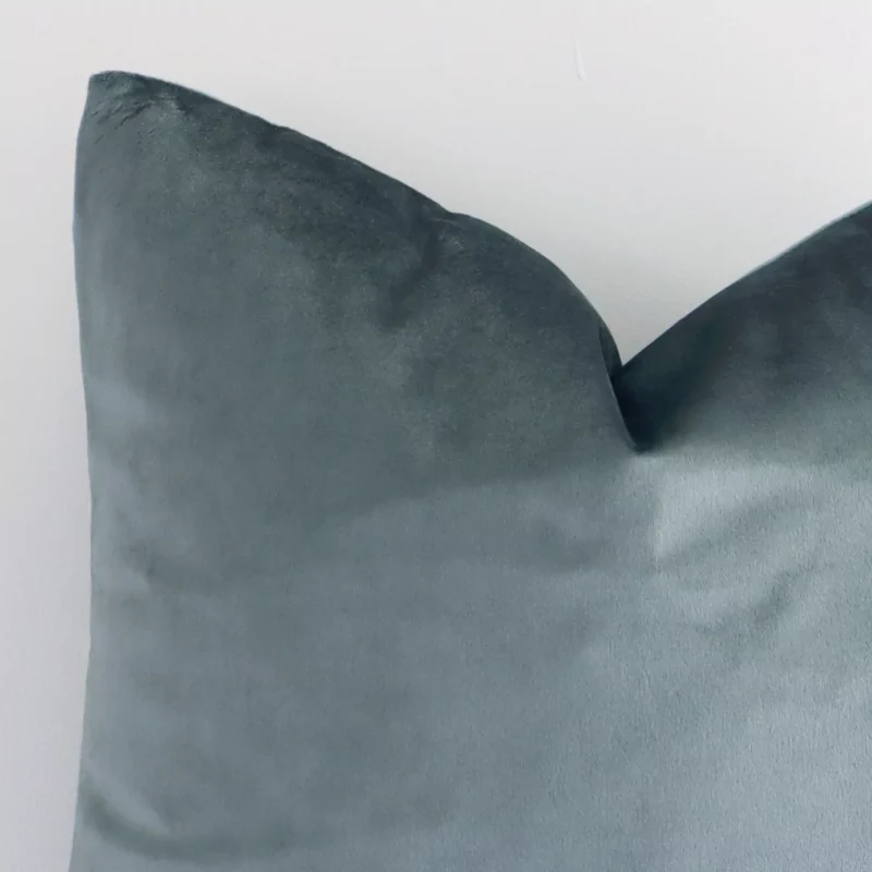 Enlarged shot of the corner of this square solid-coloured cushion cover in sage green colour is shown against a brick wall. The image shows the quality and craftsmanship of the velvet material.