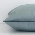 A sage green cushion arranged sideways in front of a wall. The square size and velvet fabric are shown and the seams are clearly visible.