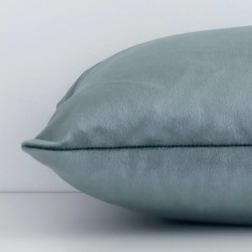 A solid-coloured sage green cushion cover is laid flat. This angle shows the side of the velvet fabric and its rectangular shape.
