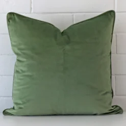 Sage cushion positioned in front of a brick wall. It has large dimensions and is made from a velvet material.