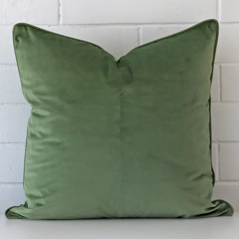 Sage cushion positioned in front of a brick wall. It has large dimensions and is made from a velvet material.